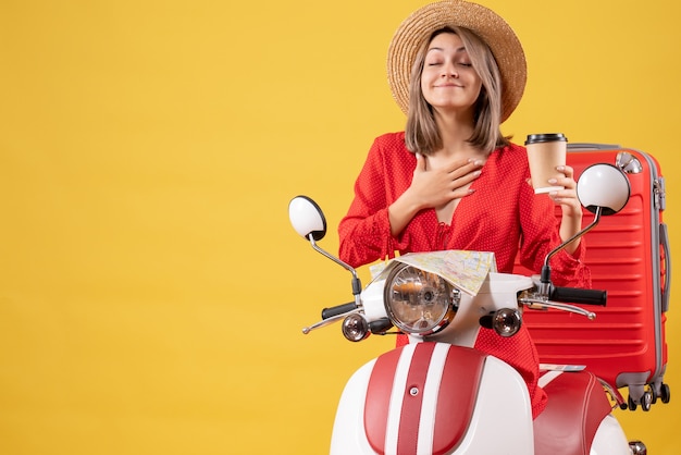 Vorderansicht junge Dame im roten Kleid, die Augen schließt, die Kaffeetasse in der Nähe von Moped hält