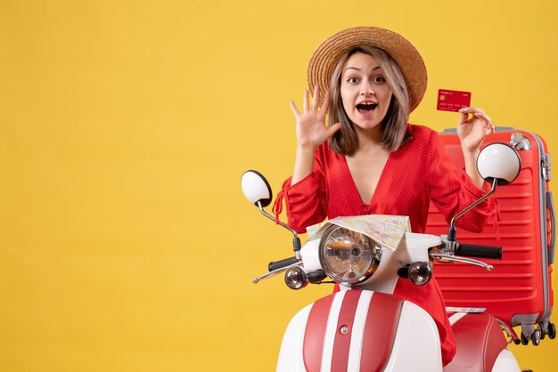 Vorderansicht junge Dame im roten Kleid mit Kreditkarte winkende Hand in der Nähe von Moped