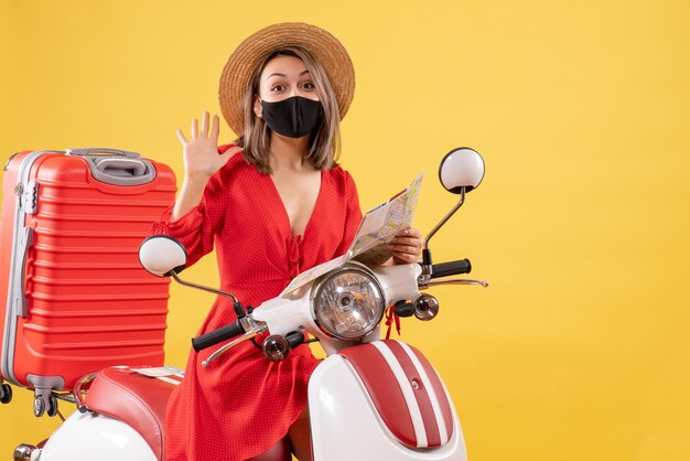 Vorderansicht junge dame mit schwarzer maske, die karte mit winkender hand in der nähe des mopeds hält