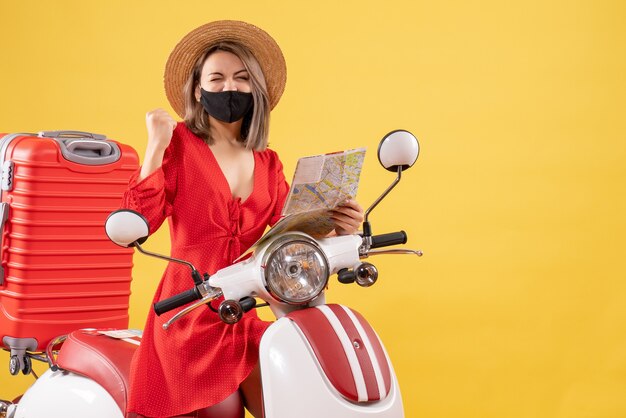 Vorderansicht junge Dame mit schwarzer Maske mit Karte, die ihr Glück in der Nähe des Mopeds ausdrückt