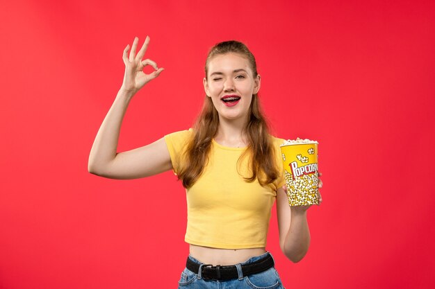 Vorderansicht junge Frau am Kino, die Popcorn-Paket hält und auf dem hellroten Wandfilmtheaterkino-Spaßfilm aufwirft