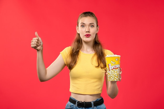 Kostenloses Foto vorderansicht junge frau am kino, die popcorn-paket hält und auf rotem wandfilmtheaterkino weiblicher spaßfilm aufwirft