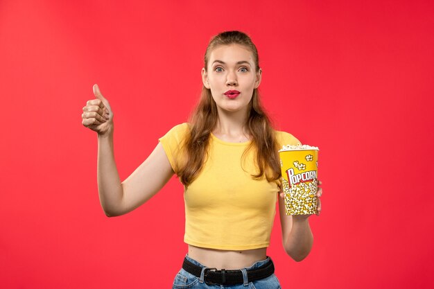 Vorderansicht junge Frau am Kino, die Popcorn-Paket hält und auf rotem Wandfilmtheaterkino weiblicher Spaßfilm aufwirft