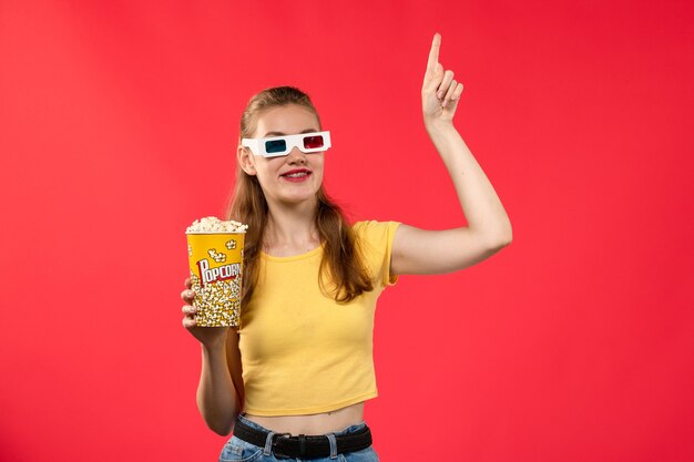 Vorderansicht junge Frau am Kino, die Popcorn-Paket in -d Sonnenbrille hält und auf rotem Wandkino-Kino-Filmmädchen lächelt