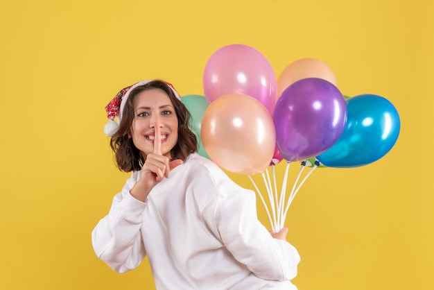 Vorderansicht junge Frau, die bunte Luftballons auf gelbem Schreibtisch Neujahrsweihnachtsfarbfeiertagsfrauenemotion versteckt