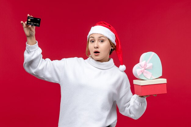 Vorderansicht junge Frau, die Geschenke und Bankkarte auf rotem Bodenfeiertag Weihnachten rot hält