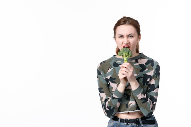 Vorderansicht junge Frau, die kleinen grünen Brokkoli hält und ihn auf weißem Hintergrund beißt