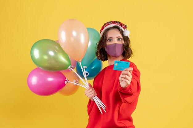 Kostenloses Foto vorderansicht junge frau, die luftballons und bankkarte in der maske auf gelb hält