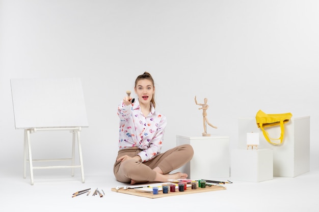 Vorderansicht junge Frau, die mit Farben sitzt und Pinsel auf weißem Hintergrund hält
