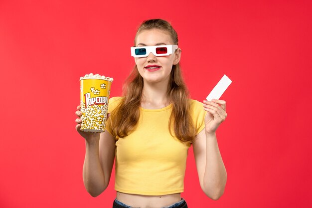 Vorderansicht junge Frau, die Popcorn-Paket und Ticket in d Sonnenbrille auf hellrotem Wandtheater-Filmkino-Film hält