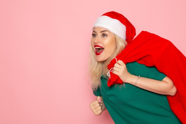 Vorderansicht junge Frau, die rote Tasche mit Geschenken auf der rosa Wandmodellfeiertagsweihnachtsfoto-Weihnachtsfärbung Santa trägt