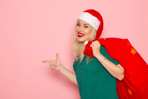 Vorderansicht junge Frau, die rote Tasche mit Geschenken auf rosa Wandfeiertagsmodell Weihnachten Neujahrsfarbe Santa trägt