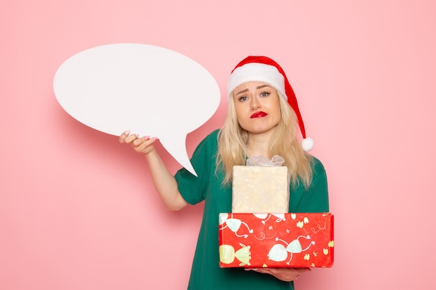 Kostenloses Foto vorderansicht junge frau hält weihnachtsgeschenke und weißes zeichen auf rosa wandfrau geschenk schnee farbfoto neujahrsfeiertag