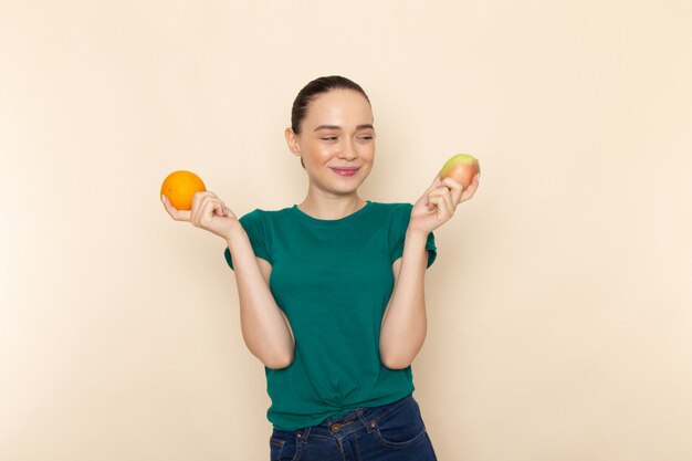Vorderansicht junge Frau im dunkelgrünen Hemd und in den blauen Jeans, die Apfel und Orange lächelnd auf Beige halten