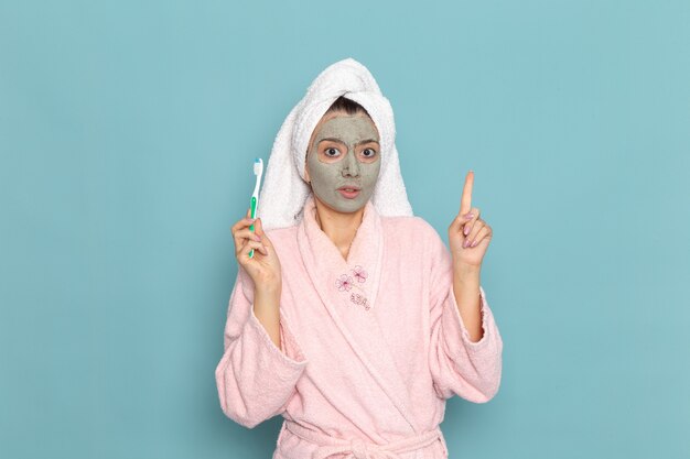 Vorderansicht junge Frau im rosa Bademantel, der Zahnbürste auf blauem Schreibtisch reinigt Schönheit Selbstpflege Creme Dusche
