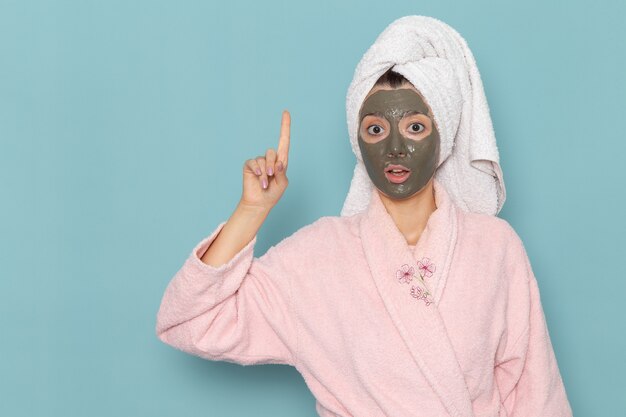 Vorderansicht junge Frau im rosa Bademantel mit Maske auf ihrem Gesicht, das auf der blauen Wandduschenreinigungsschönheitselbstpflegecreme aufwirft
