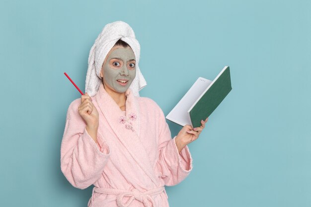 Vorderansicht junge Frau im rosa Bademantel mit Maske auf ihrem Gesicht, das Heft auf blauer Wanddusche reinigt, die Schönheits-Selbstpflegecreme reinigt