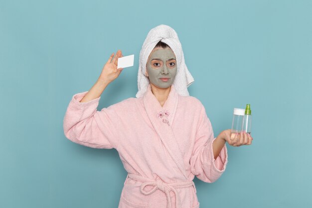 Vorderansicht junge Frau im rosa Bademantel nach Dusche, die Spray und Karte auf der blauen Wandschönheit Wasserbadcreme Selbstpflege-Duschbad hält