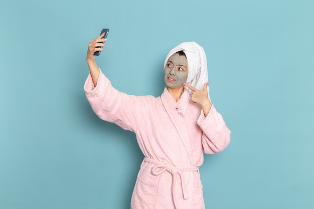 Vorderansicht junge Frau im rosa Bademantel unter Selfie mit Maske auf ihrem Gesicht auf blauem Schreibtisch, der Schönheits-Selbstpflege-Cremedusche reinigt