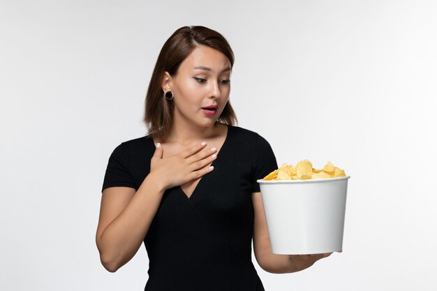 Vorderansicht junge Frau im schwarzen Hemd, das Kartoffelchips hält und auf hellweißer Oberfläche aufwirft