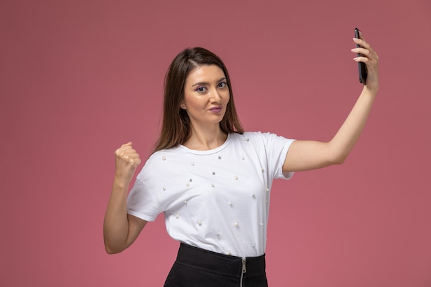 Vorderansicht junge Frau im weißen Hemd, das ein Selfie auf der rosa Wand, Modellfrau nimmt