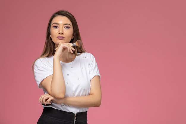 Vorderansicht junge Frau im weißen Hemd, das Make-up-Pinsel auf der rosa Wand hält, Fotofarbenfrau-Posenmodell