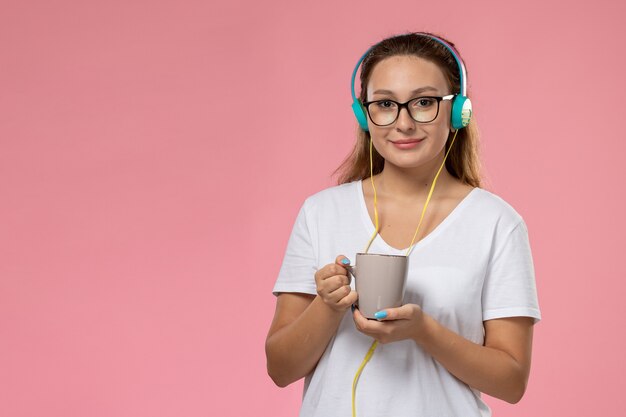 Vorderansicht junge Frau im weißen T-Shirt, das gerade Musik über Kopfhörer hört und Tasse mit Tee auf dem rosa Hintergrund hält