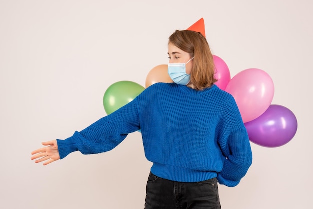 Vorderansicht junge Frau in der sterilen Maske, die bunte Luftballons hinter ihrem Rücken versteckt