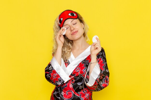Vorderansicht junge Frau in Nachtgewand und tragende Augenmaske, die ihr Gesicht auf gelbem Wandschlafmädchen-Nachtbettmodell reinigt
