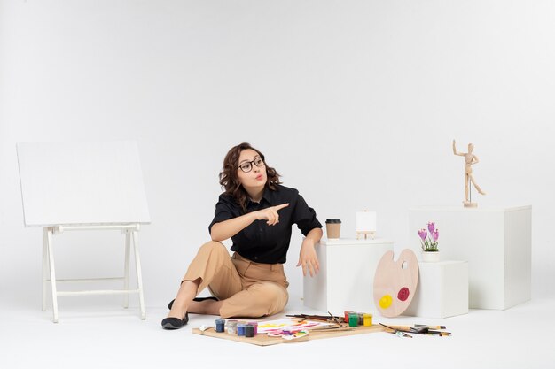 Vorderansicht junge Frau sitzt im Zimmer mit Staffelei und Farben auf weißem Hintergrund