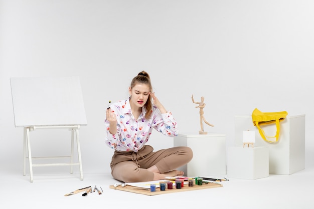 Vorderansicht junge Frau sitzt mit Farben und Staffelei zum Zeichnen auf weißem Hintergrund
