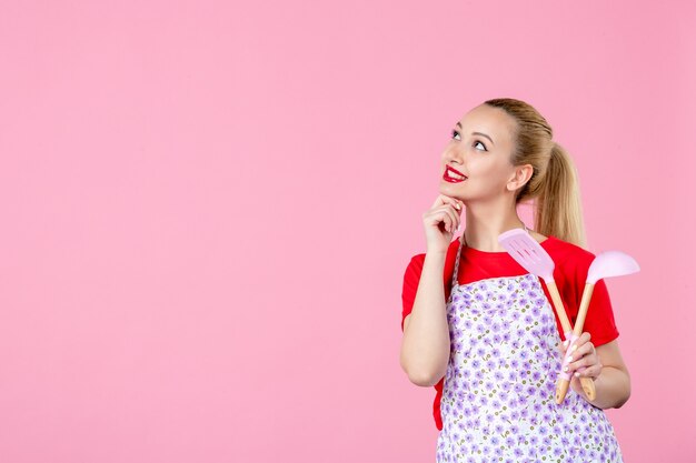 Vorderansicht junge Hausfrau im Umhang mit Besteck an rosa Wand