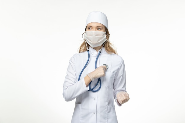 Vorderansicht junge Ärztin im medizinischen Anzug, die Schutzmaske wegen Coronavirus unter Verwendung des Stethoskops auf der hellweißen Oberfläche trägt