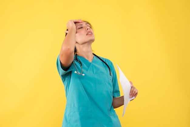 Vorderansicht junge Ärztin im medizinischen Hemd, das Papieranalyse hält, betont auf gelbem Hintergrund