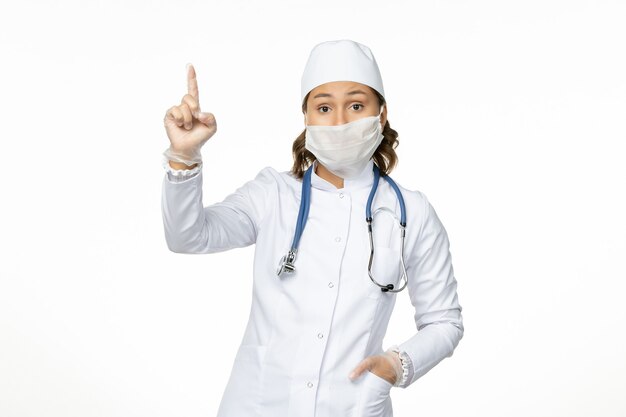 Vorderansicht junge Ärztin im weißen medizinischen Anzug und mit Maske wegen Coronavirus auf dem hellweißen Schreibtisch