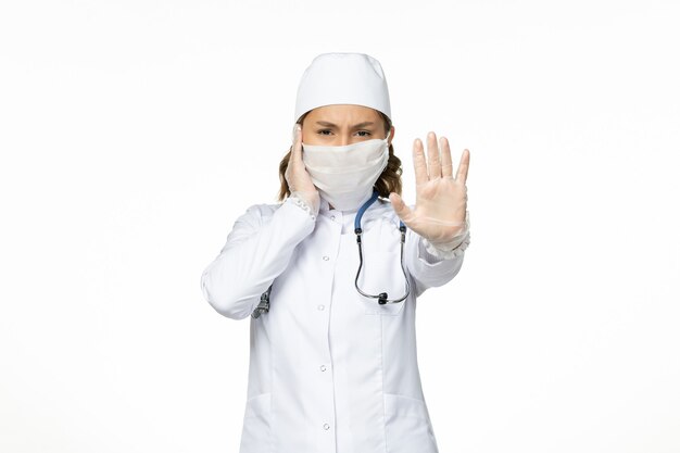 Vorderansicht junge Ärztin im weißen medizinischen Anzug und mit steriler Maske wegen Coronavirus auf weißem Schreibtisch