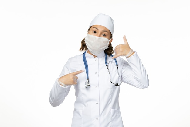 Vorderansicht junge Ärztin im weißen medizinischen Anzug und mit steriler Maske wegen Coronavirus auf weißer Oberfläche