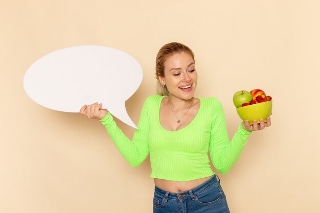 Vorderansicht junge schöne Frau im grünen Hemd, das Platte voll von Früchten mit einem großen weißen Zeichen auf Cremewandfruchtmodellfrau hält