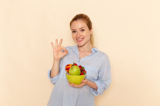 Vorderansicht junge schöne Frau im Hemd, das Platte mit Früchten anlächelt, die auf der cremefarbenen Wandfrucht-reifen Modellfrauenhaltung lächeln