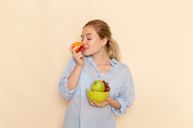 Vorderansicht junge schöne Frau im Hemd, das Platte mit Früchten hält, die Apfel auf der cremefarbenen Wandfrucht reifen Modellfrauenhaltung riechen