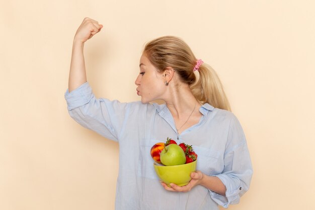 Vorderansicht junge schöne Frau im Hemd, das Platte mit Früchten hält und auf der cremefarbenen Wandfrucht reife Modellfrau posiert