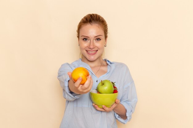 Vorderansicht junge schöne Frau im Hemd, das Platte mit Früchten mit einem leichten Lächeln auf der cremefarbenen Wandfrucht reife Modellfrauenhaltung hält