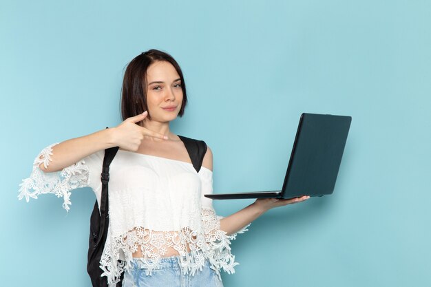 Vorderansicht junge Studentin in weißen Hemdblaujeans und schwarzer Tasche unter Verwendung des Laptops auf dem blauen Raum der weiblichen Studentenuniversitätsschule