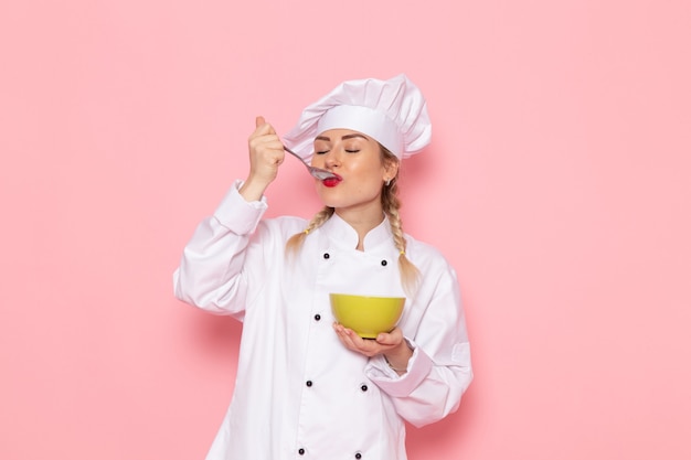 Vorderansicht junge weibliche Köchin im weißen Kochanzug, der Suppe auf dem Arbeitsfoto der rosa Raumkochküche schmeckt