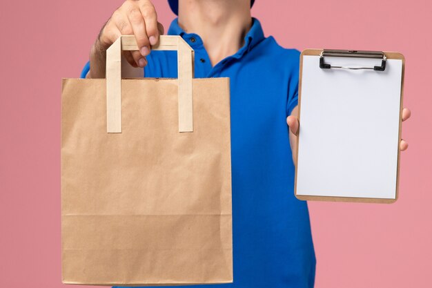 Vorderansicht junger männlicher Kurier in blauer Uniform und Umhang mit Lieferpaket und Notizblock auf seinen Händen an der rosa Wand