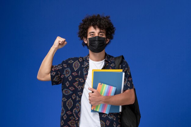 Vorderansicht junger männlicher Student, der schwarze Maske mit Rucksack hält, der Heft hält und Dateien, die sich auf blauem Hintergrund freuen.