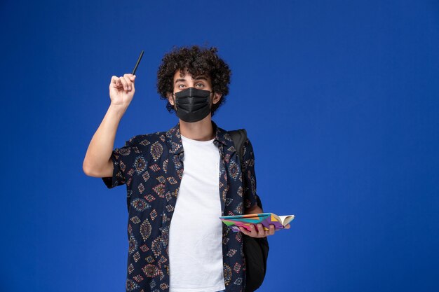 Vorderansicht junger männlicher Student, der schwarze Maske mit Rucksack hält, der Heft und Stift auf blauem Schreibtisch hält.