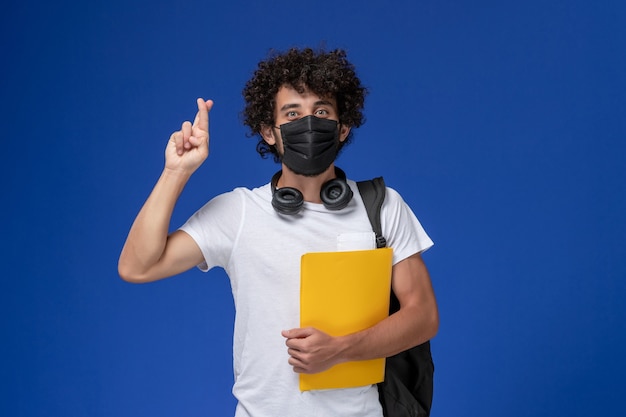 Vorderansicht junger männlicher Student im weißen T-Shirt, das schwarze Maske trägt und gelbe Akten auf dem hellblauen Schreibtisch hält.