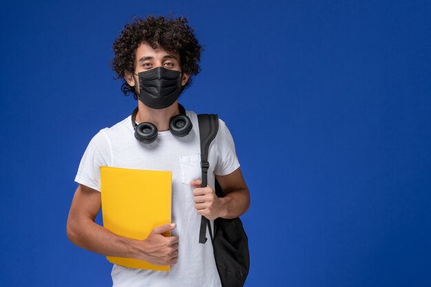 Vorderansicht junger männlicher Student im weißen T-Shirt, das schwarze Maske trägt und gelbe Dateien auf dem hellblauen Hintergrund hält.