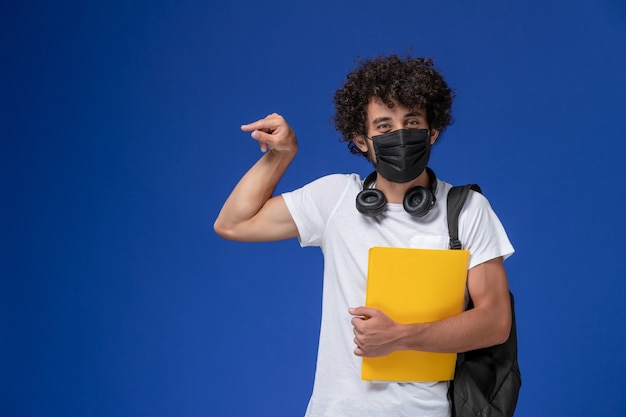 Vorderansicht junger männlicher Student im weißen T-Shirt, das schwarze Maske trägt und gelbe Dateien auf dem hellblauen Hintergrund hält.